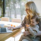 A mother works on a laptop at home while holding her baby, showcasing remote working and parenting.