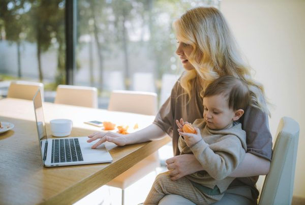 A mother works on a laptop at home while holding her baby, showcasing remote working and parenting.
