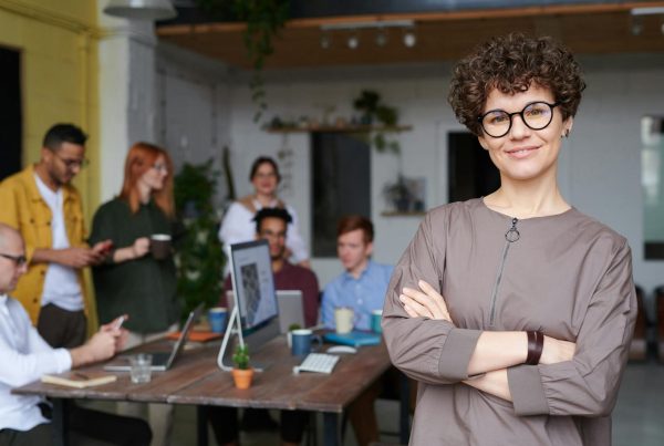 Smiling businesswoman with curly hair stands confidently in a modern office space with colleagues.