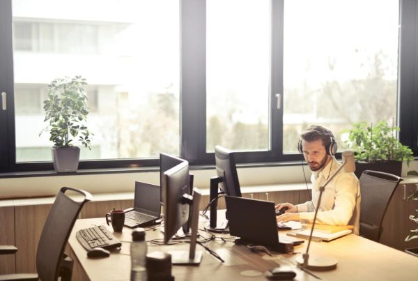 A businessman sits at a desk using multiple computers and a headset in a well-lit modern office.