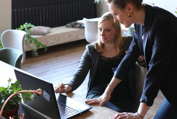 Two professional women discuss a project using a laptop in a modern office environment.