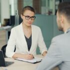 Woman in glasses interviews man at office desk.