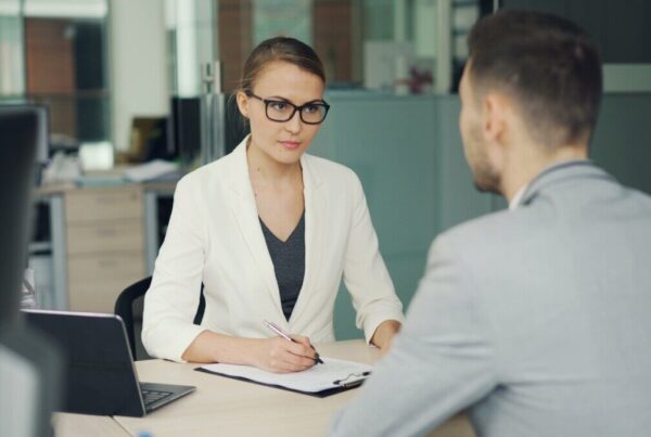 Woman in glasses interviews man at office desk.